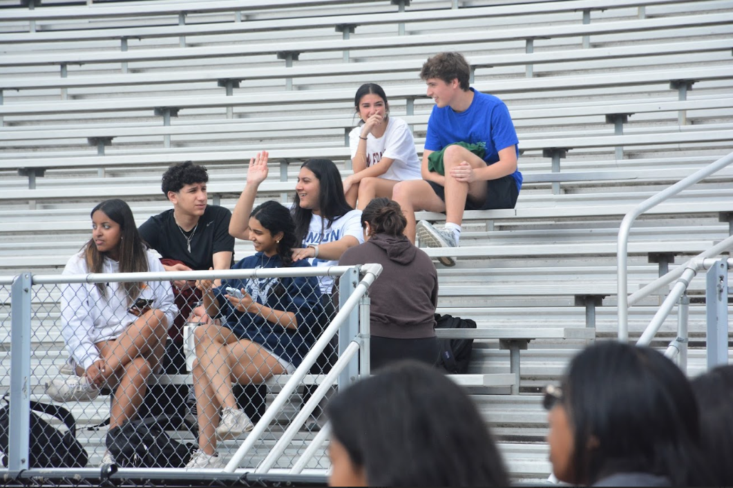 Students chat and watch others playing on the field from the bleachers.